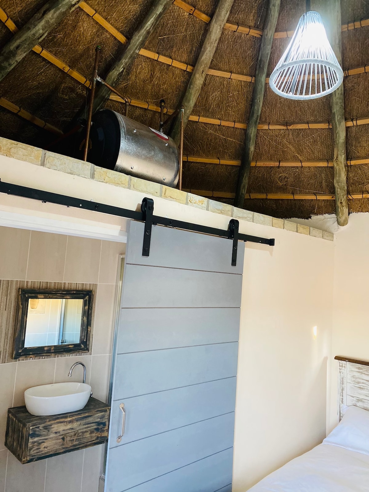 A uniquely styled room features a circular thatched ceiling supported by wooden beams. A sliding barn door leads to a compact bathroom. A minimalist basin sits on a rustic shelf. A pendant light hangs above, adding brightness to the space.