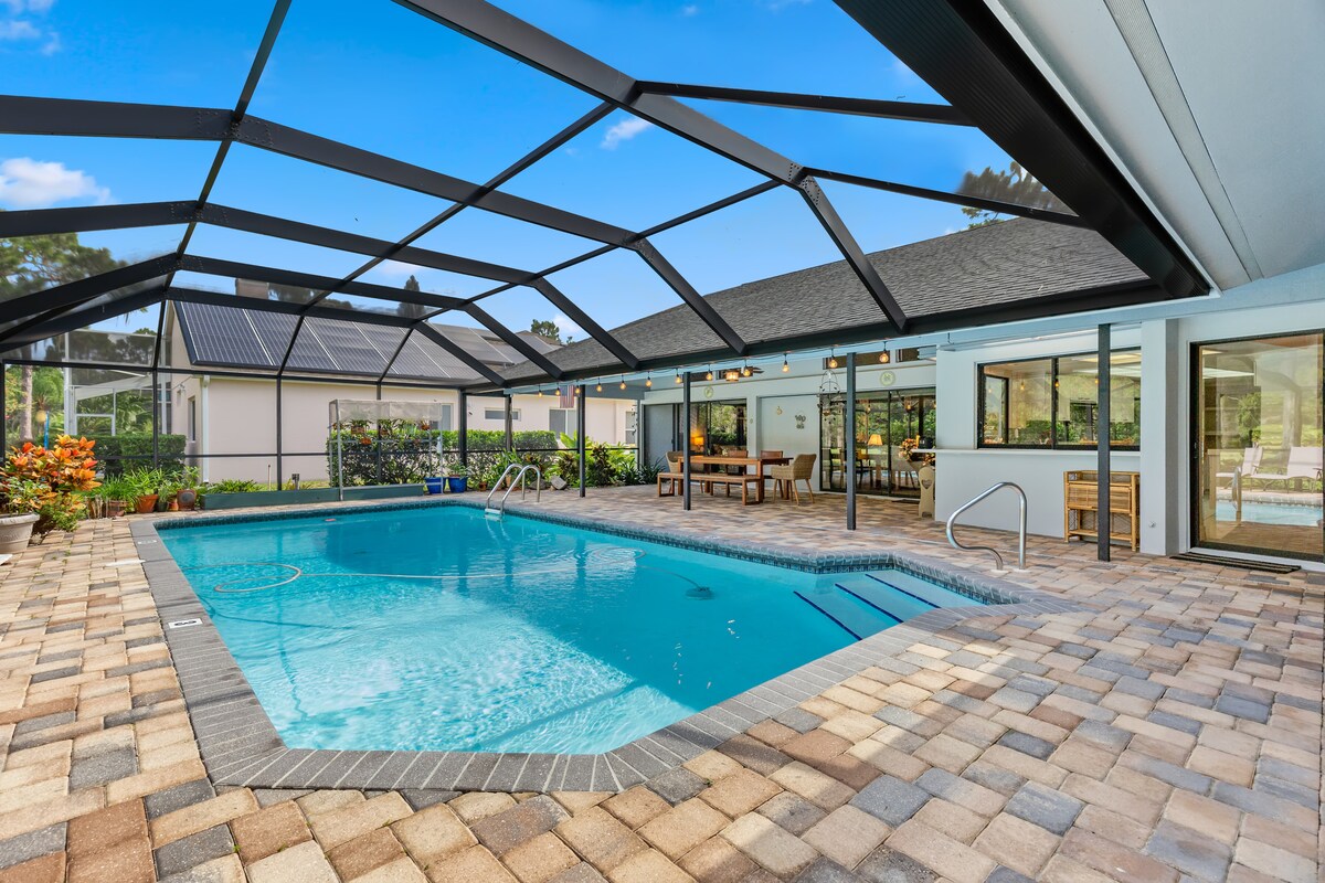 A spacious pool area is enclosed with a screen for comfort. The crystal-clear water is surrounded by paver stones, with lounge chairs positioned for relaxation. Nearby, dining space is visible, complemented by vibrant landscaping in the background, enhancing the serene atmosphere.