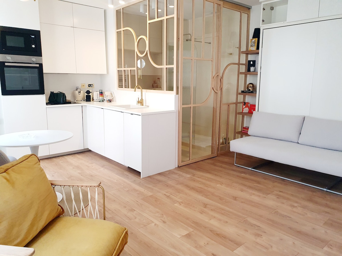A modern living space features a light wood floor and a mix of white cabinetry and decorative glass partitions. A cozy gray sofa and a yellow accent chair are present, complemented by a round dining table. Kitchen appliances and storage are seamlessly integrated.