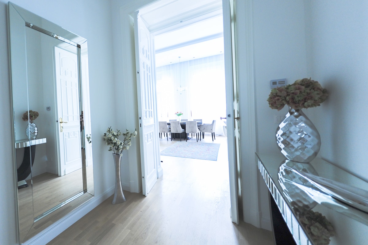 A bright entrance showcases a large mirror, a decorative vase with flowers, and a sleek console table. Light fills the space from the adjoining dining area, visible through open double doors. Neutral tones and wooden flooring contribute to a modern yet elegant aesthetic.