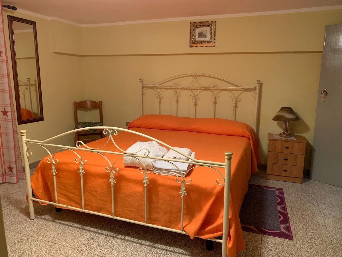 A well-furnished sleeping area features an ornate metal bed dressed in vibrant orange linens. A wooden chair sits beside a nightstand with a lamp. A mirror reflects the space, while light filters in from a window, contributing to the room's welcoming simplicity.