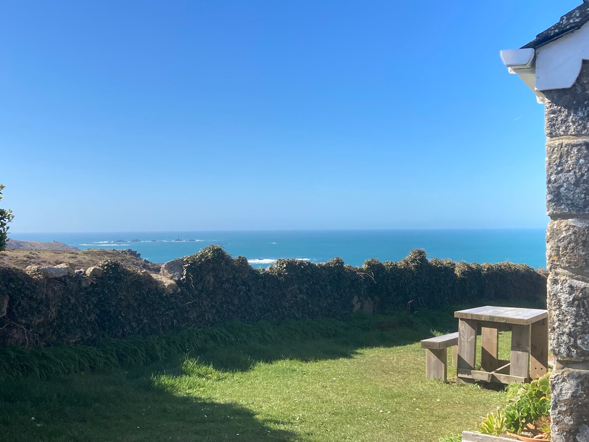 A view from the cottage shows a lush green garden leading to the cliff edge, where the vibrant blue sea is visible under a clear sky. A wooden table is positioned in the garden, offering a space to enjoy the serene coastal scenery.