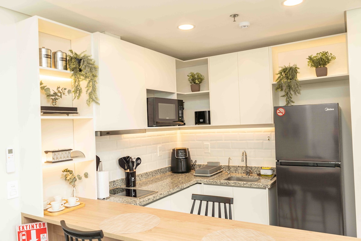 The kitchen area features modern cabinetry with open shelves displaying decorative plants. A countertop is equipped with essential appliances, including a microwave and coffee maker, along with a spacious fridge. The harmonious blend of functionality and style is enhanced by soft lighting.