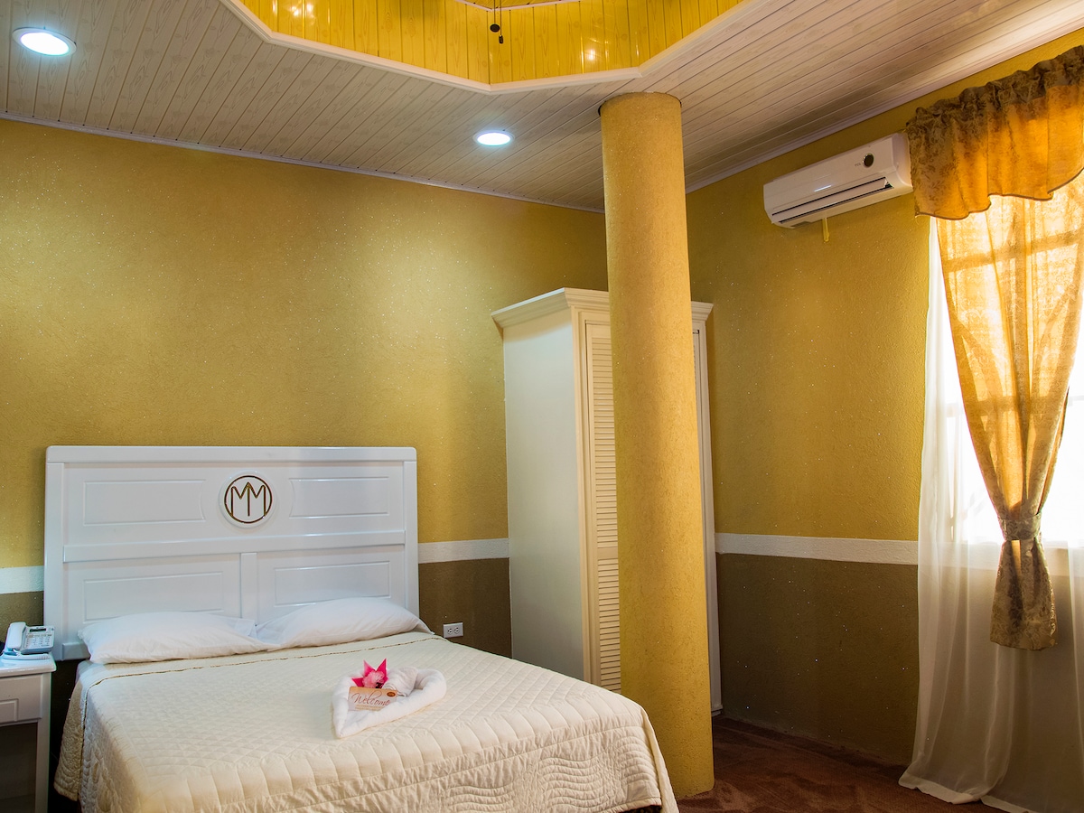A bright guest room features a spacious bed with a light-patterned quilt. Soft golden walls complement the warm lighting. An air conditioning unit is mounted above an elegant window dressed with sheer curtains, allowing natural light to enter. A tall wardrobe is positioned nearby.