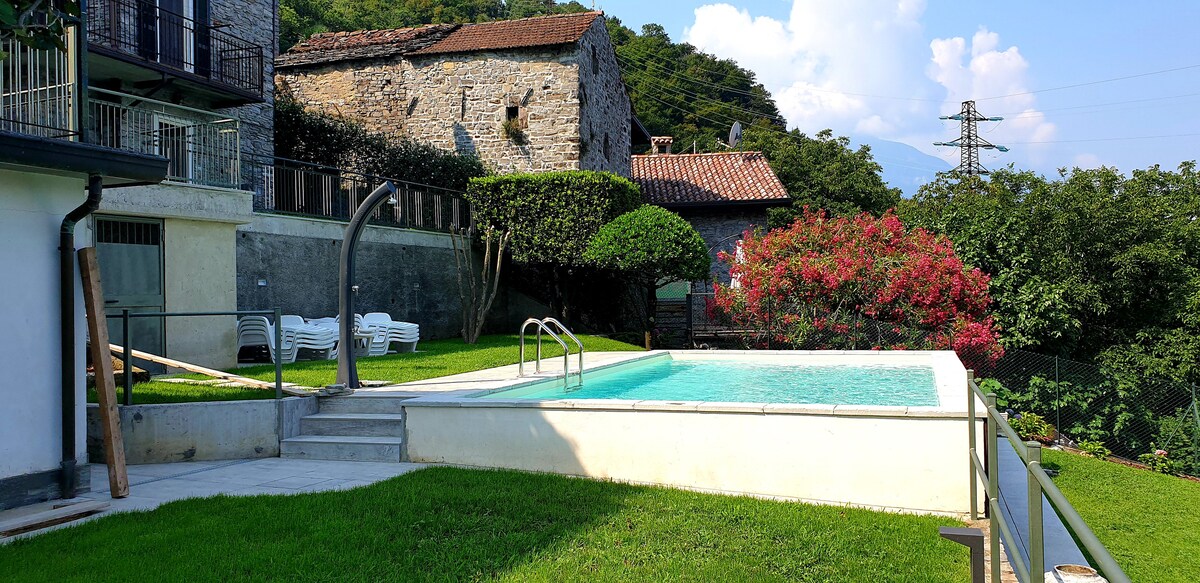 A tranquil outdoor space features a swimming pool bordered by green grass and flowering plants. The pool is equipped with a metal ladder, and several lounge chairs are positioned nearby. Stone buildings are visible in the background, contributing to the rustic ambiance.