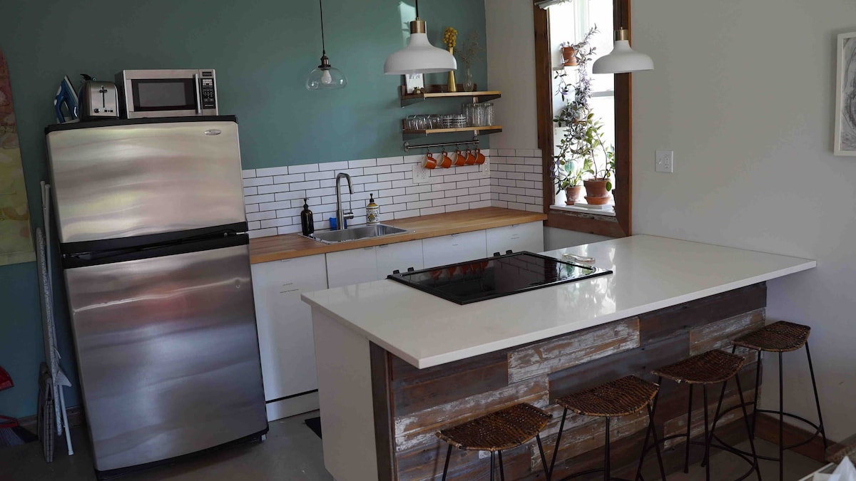 A modern kitchen features stainless steel appliances, including a refrigerator and microwave. A white countertop surrounds a cooktop, with bar stools positioned at the island. Open shelving displays glassware and orange accents, while natural light filters in through the window, illuminating the space.