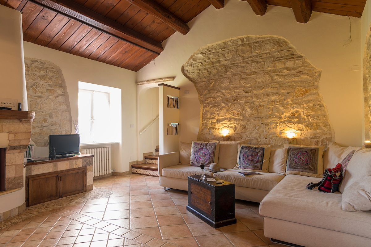 A living room features a cozy arrangement of a sectional sofa with decorative cushions. Exposed wooden beams and natural stone walls create a warm ambiance. A small TV sits on a wooden cabinet, while a window allows natural light to fill the space, adjacent to a staircase.