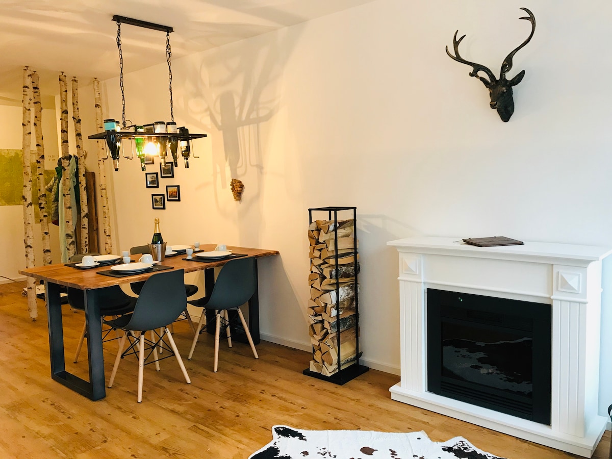 A dining area features a wooden table set for four, complemented by modern chairs. A decorative fireplace adds warmth, while a stylish log holder visually enhances the space. The walls are adorned with artwork, and shadow patterns from hanging light fixtures create an inviting atmosphere.
