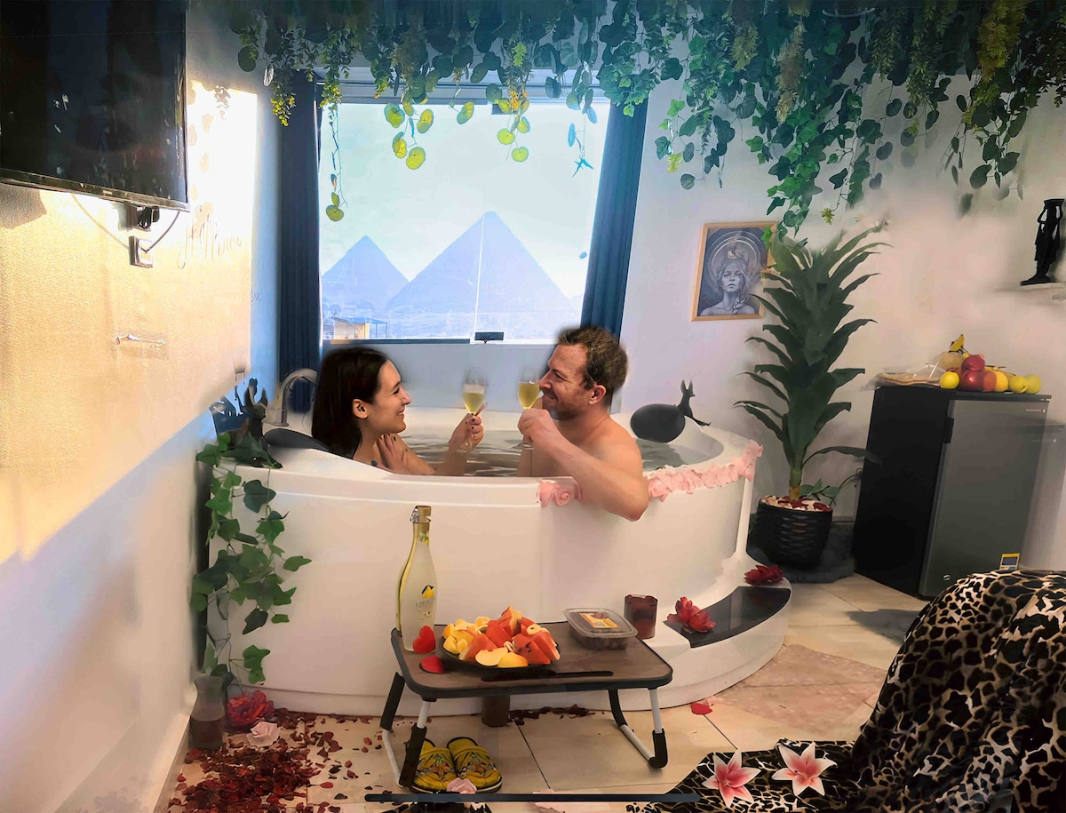 A luxurious soaking tub is surrounded by lush greenery and decorative flowers. A small table holds refreshments, while the backdrop features large windows showcasing a view. Soft lighting creates a serene ambiance, with artistic elements adding to the overall decor.