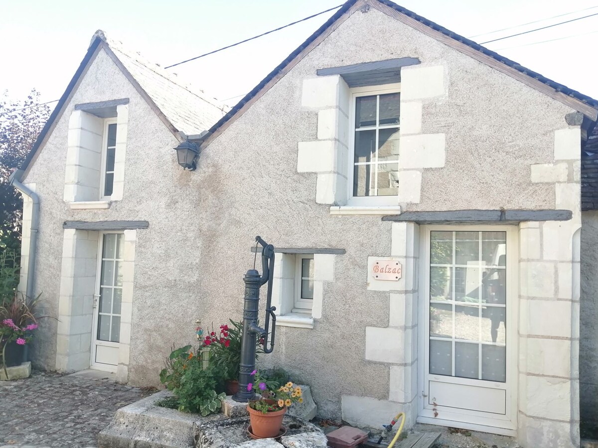 The exterior of a charming stone cottage features a textured facade with white trim. An antique water pump is positioned in front, surrounded by colorful flowers in a pot. Large windows with shutters enhance the inviting appearance of the entrance.
