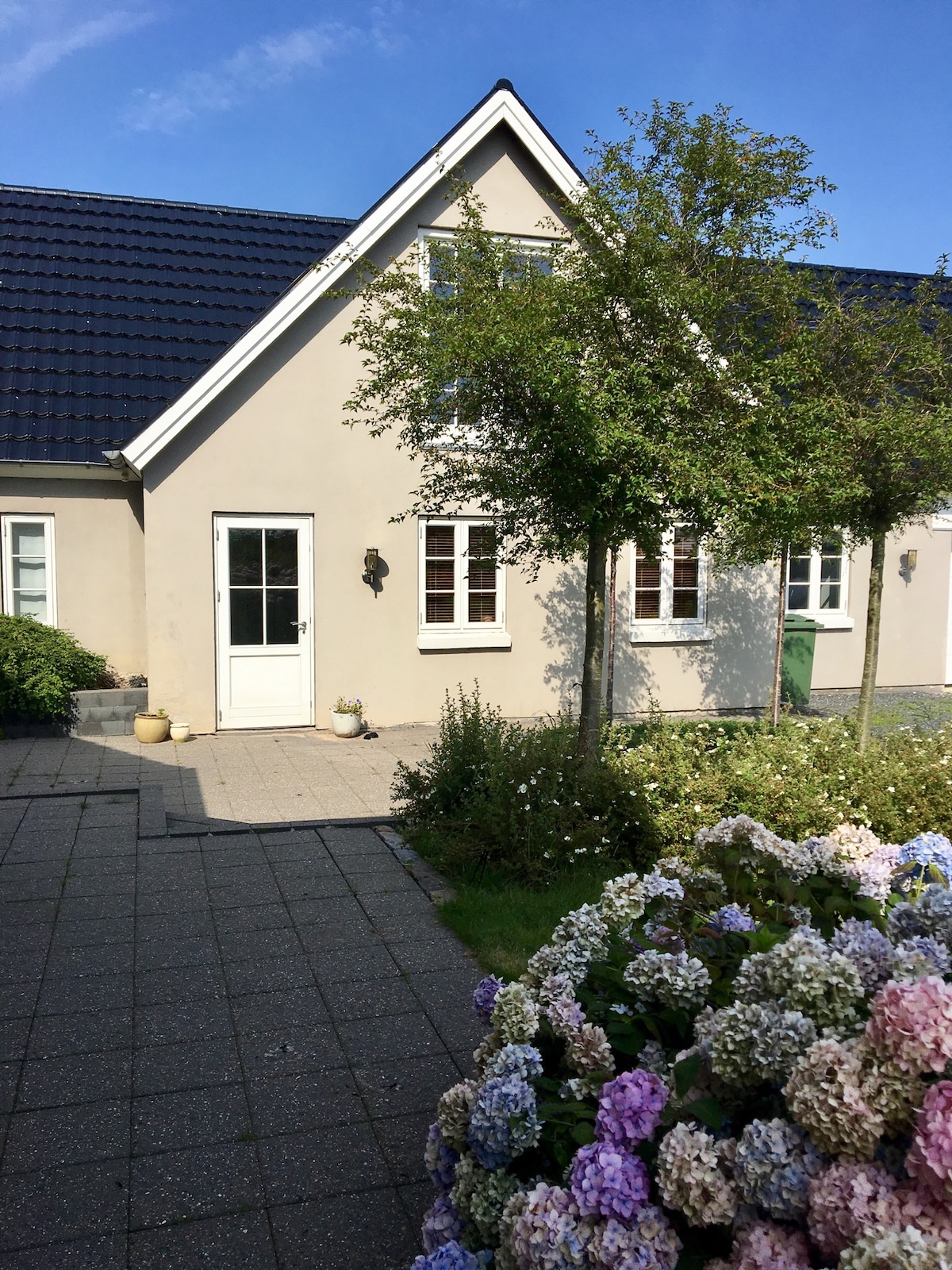 A welcoming exterior of a charming building is showcased, featuring a light-colored façade and a sloped roof. The entrance is highlighted by a white door and flanked by several small windows. Colorful flowers in the foreground add a natural touch to the landscape.