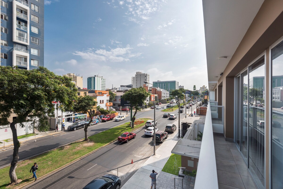 A balcony view overlooks a busy street lined with trees, showcasing the vibrant atmosphere of the area. Modern buildings and vehicles are visible in the distance, while pedestrians walk along the sidewalk. Natural light fills the outdoor space, highlighting the welcoming environment.