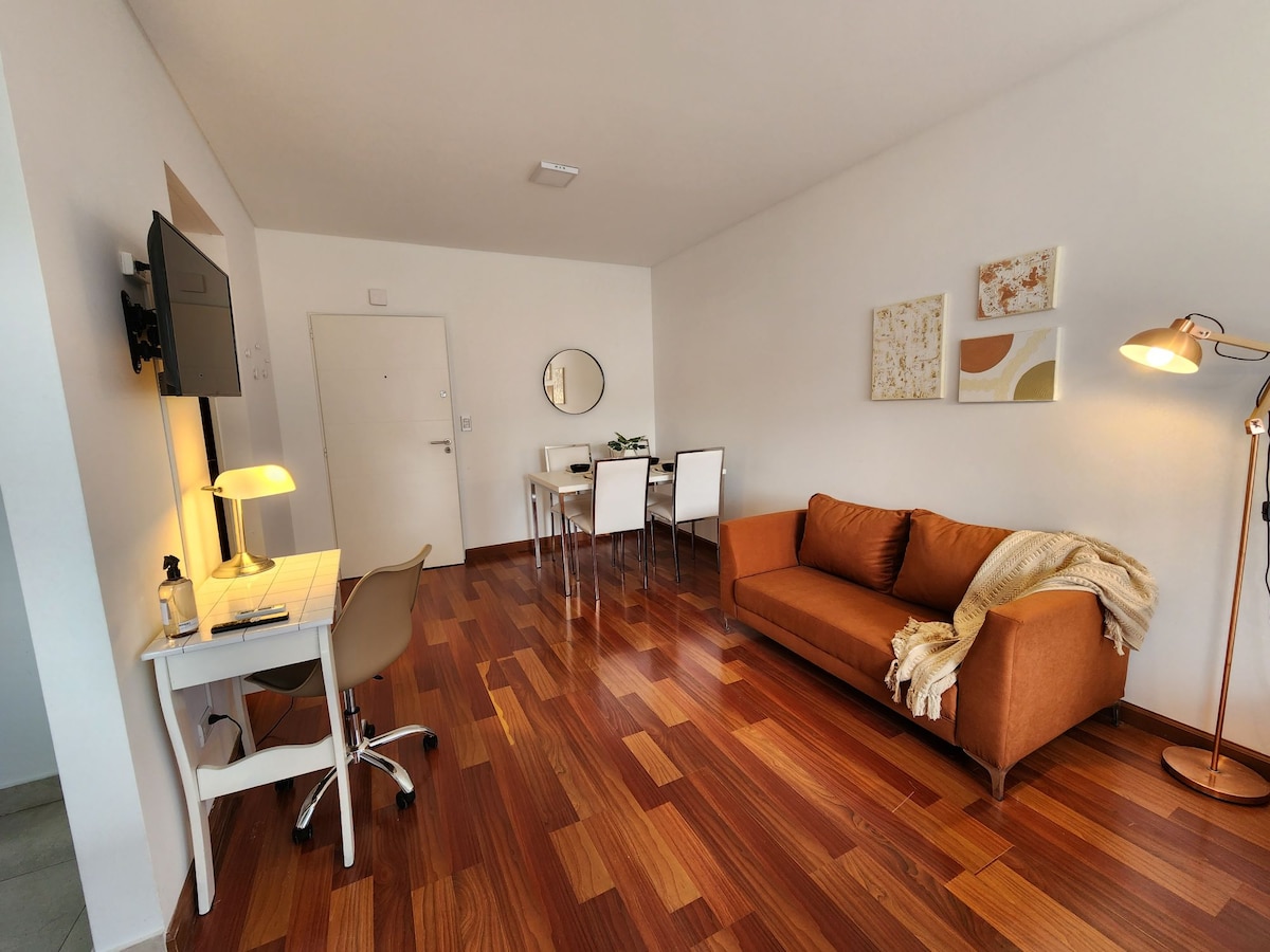 A sunlit living area features hardwood flooring, providing a warm ambiance. A brown couch is complemented by a textured throw. A small desk with a lamp is situated beside a wall-mounted TV. A dining table for four is positioned in the background, against a white wall with decorative artwork.