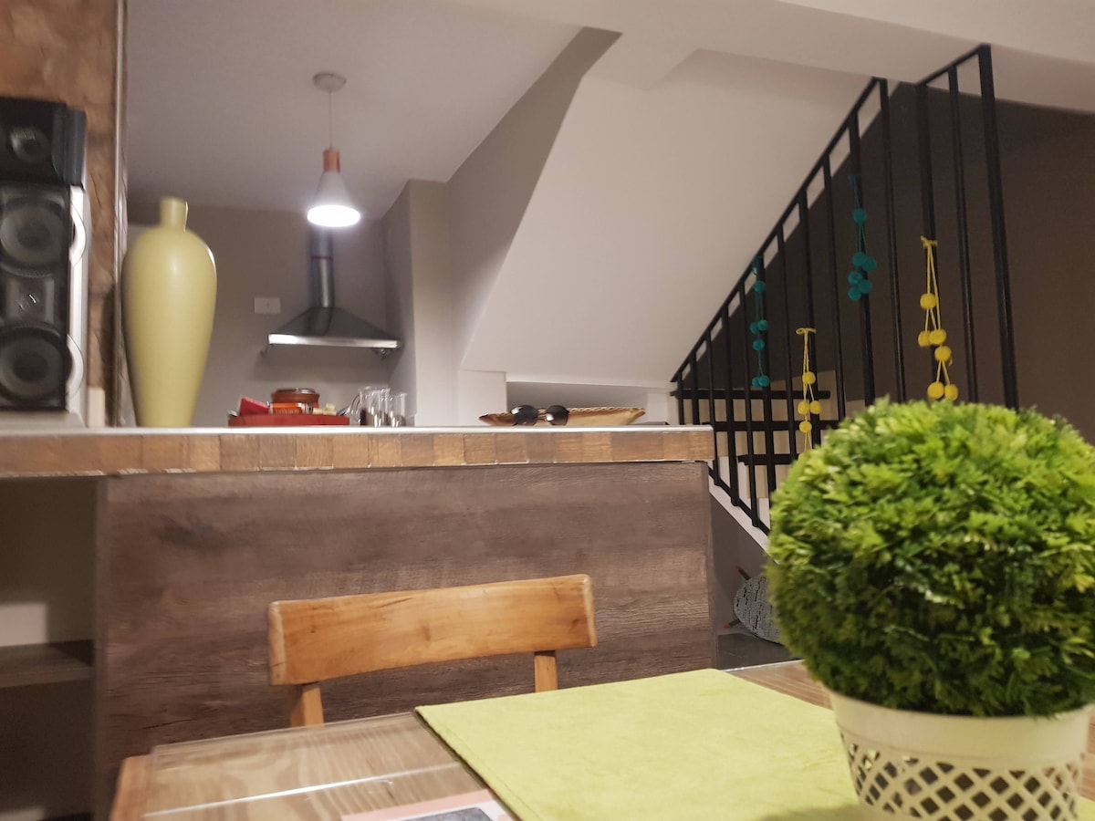 A wooden dining table is set with a green placemat, complemented by a small decorative potted plant. In the background, a staircase ascends, while a slim kitchen area is visible, featuring a hanging light fixture and various kitchen utensils. Two audio speakers are positioned on a counter.