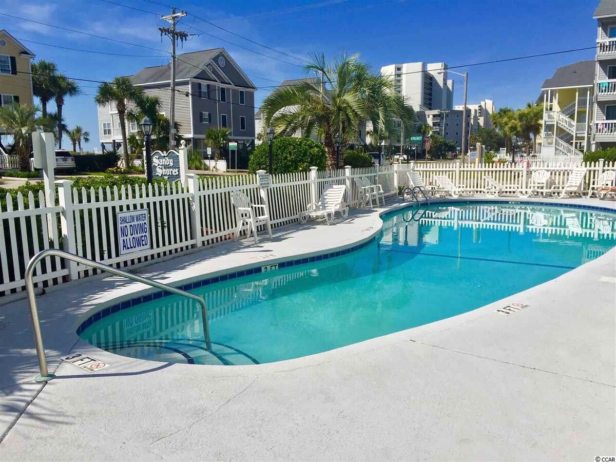 A serene swimming pool is presented, surrounded by a white picket fence and well-maintained landscaping. Several lounge chairs are neatly arranged around the pool area, and palm trees provide a touch of greenery against a clear blue sky.