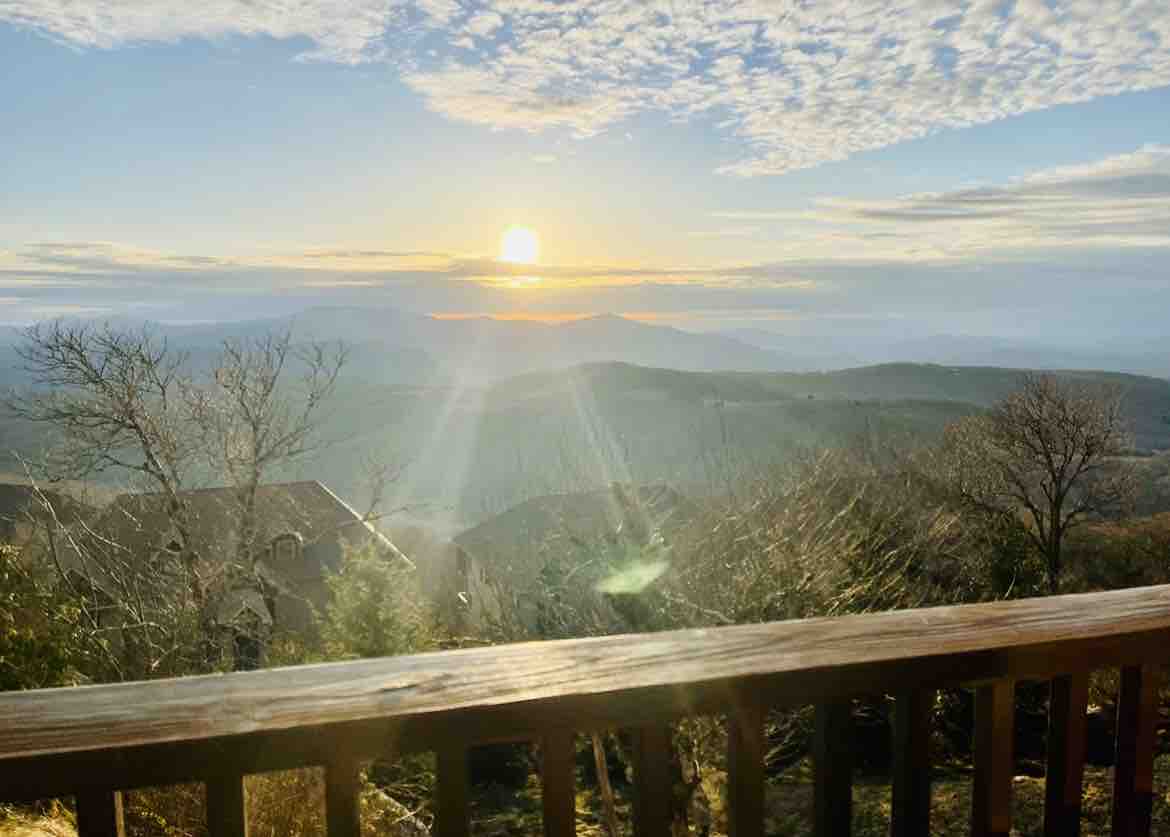 A wooden railing frames a panoramic view of the Blue Ridge Mountains at sunset. The sun casts a warm glow over the landscape, highlighting the gentle slopes and tree silhouettes. A serene atmosphere is established, inviting appreciation of the natural beauty.