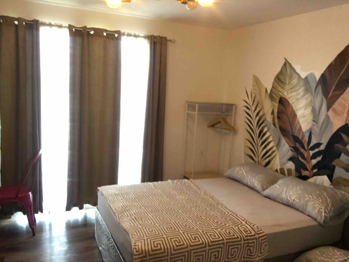 A spacious studio room features a double-sized bed with soft bedding and a decorative geometric blanket. Light floods in through large windows dressed with curtain panels. A small closet with hangers and a bright red chair add character to the cozy atmosphere.