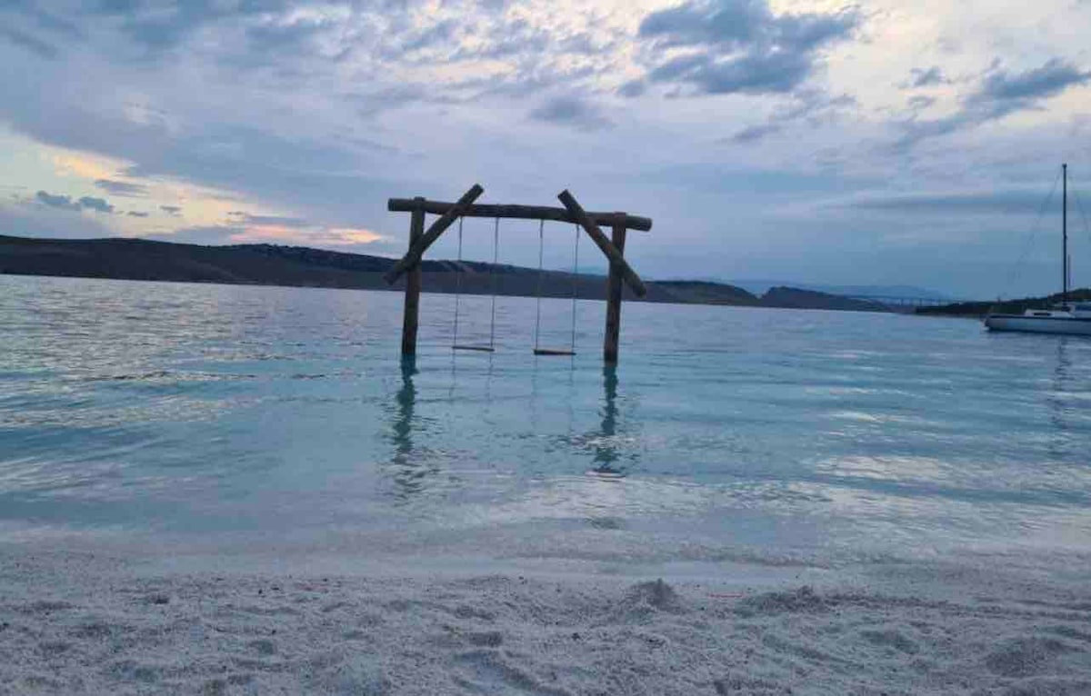 A wooden swing is suspended from a simple frame, resting just above calm waters. Soft white sand lines the shore, with gentle waves lapping nearby. A sailboat is visible in the distance against a backdrop of muted clouds and a fading sky.