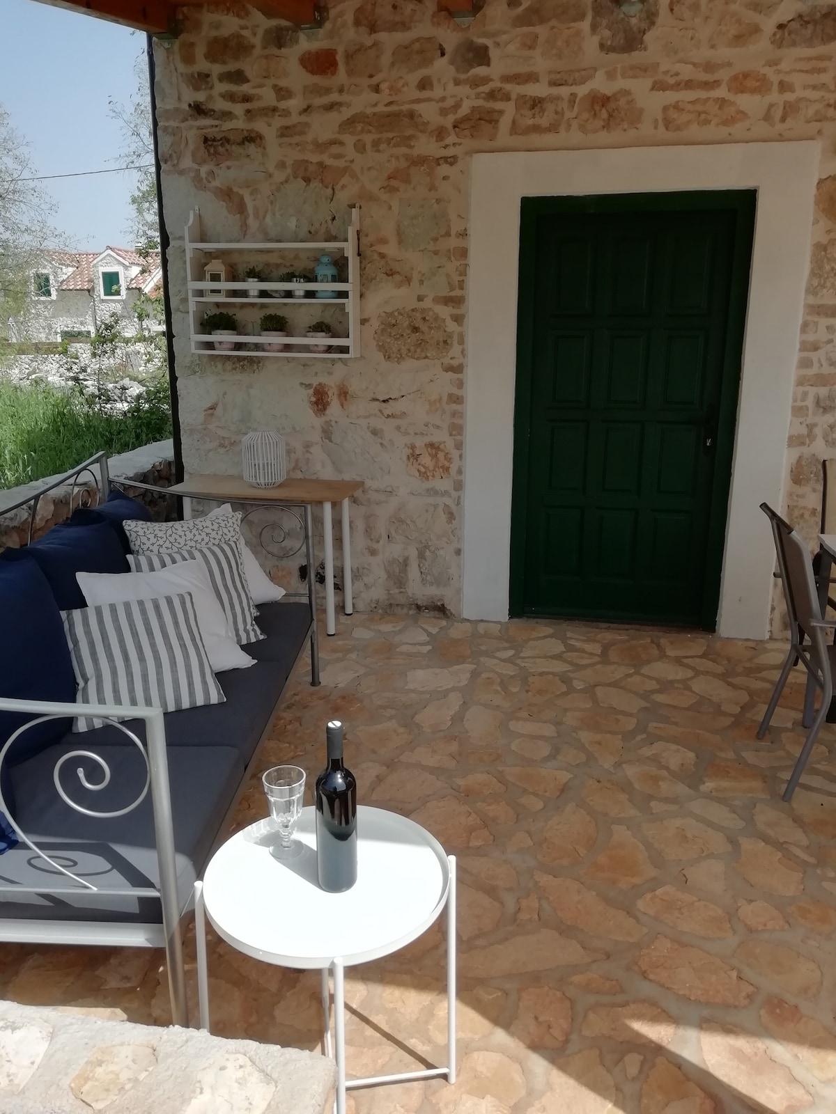 An outdoor seating area features a dark blue sofa with decorative pillows, positioned on a stone floor. A small white table holds a glass and a bottle of wine. A green door provides access to the interior, while shelves display potted plants in the background.