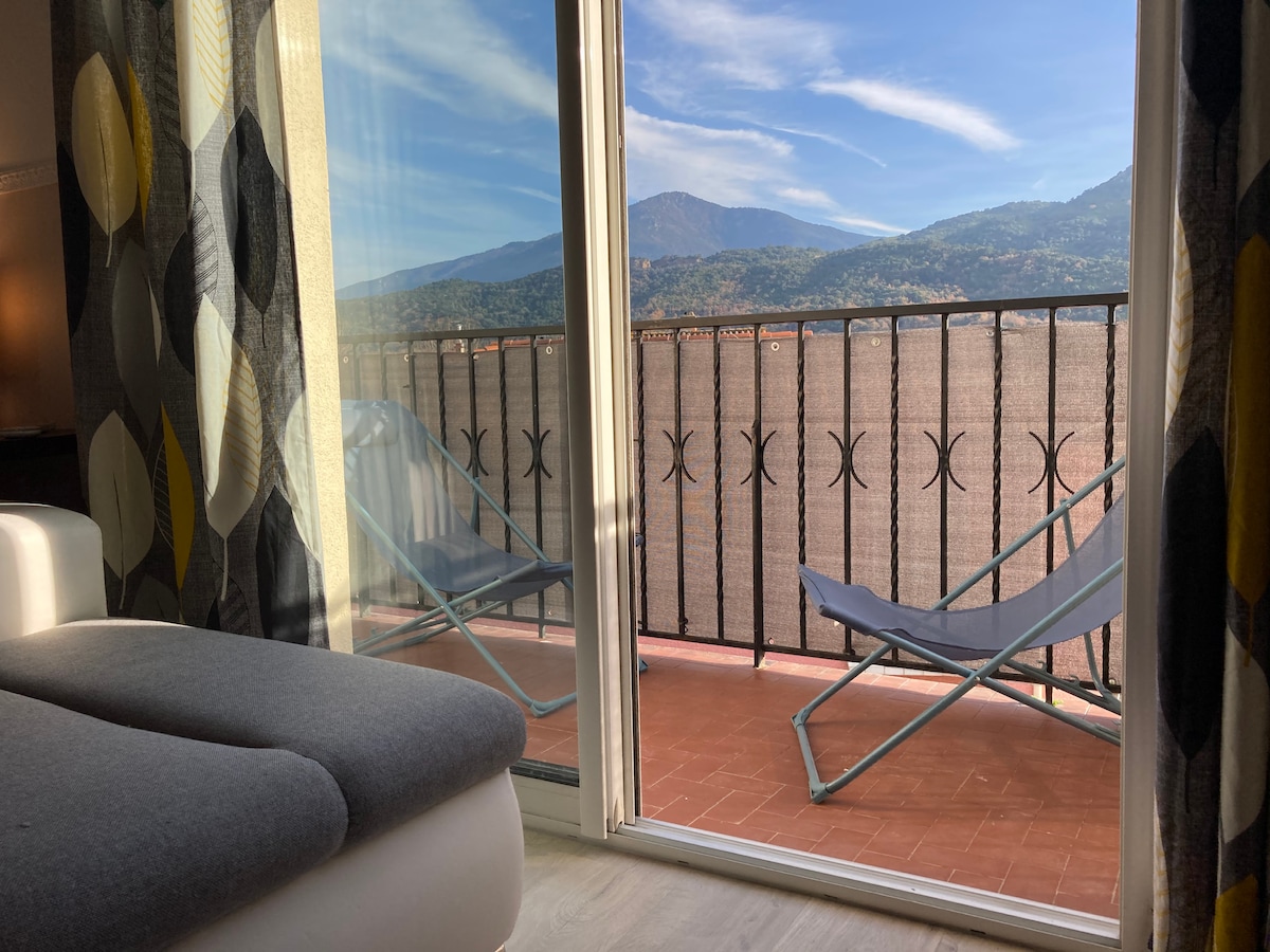 A bright and airy living space is captured, showcasing large glass doors that open to a spacious balcony. The view features distant mountains under a clear sky, while two lounge chairs are positioned on the balcony for comfortable outdoor relaxation.