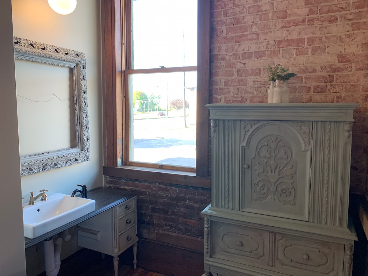 A charming bathroom area features exposed brick walls and a large window offering natural light. An antique-style blue cabinet stands beside a wooden vanity with a sink and modern fixtures. An ornate decorative mirror is mounted on the wall, providing a vintage touch.