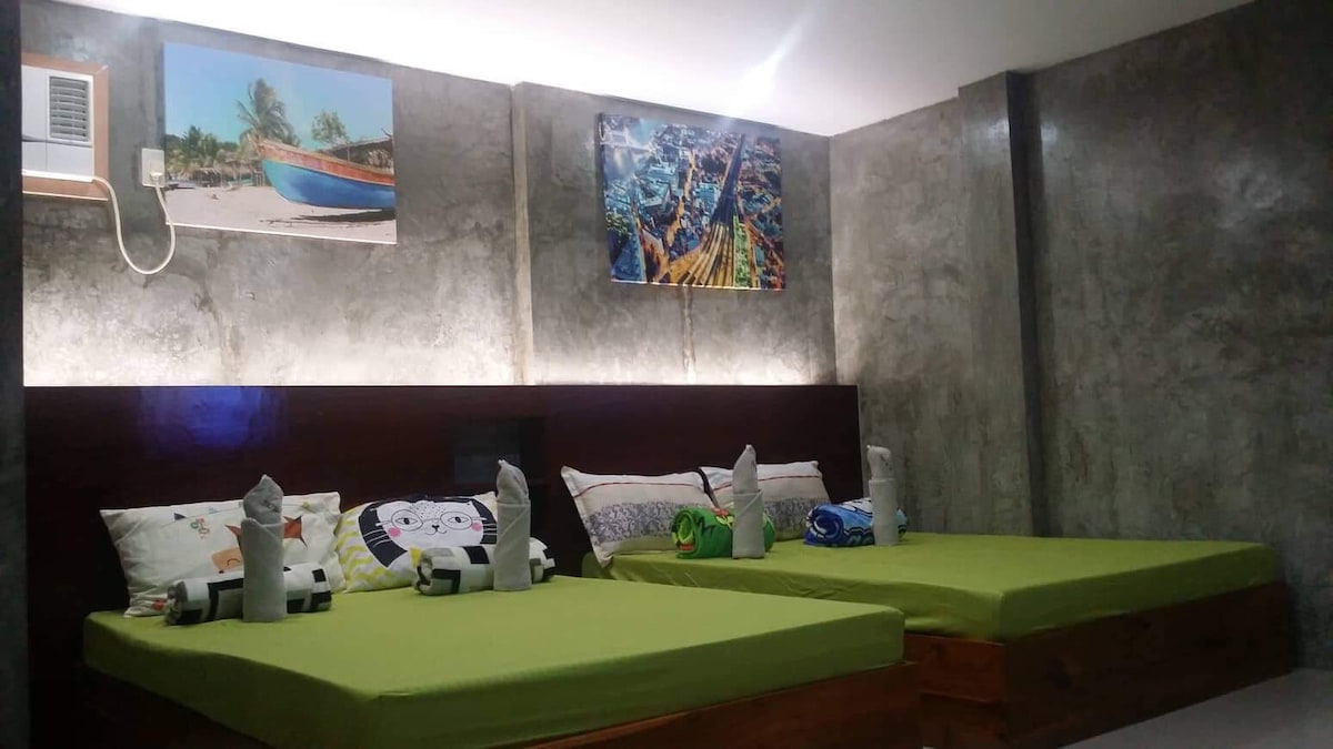 The room features two spacious beds dressed in green bedding. Decorative pillows are arranged on each bed, complemented by mounted artwork on the walls. Soft lighting provides a gentle glow, enhancing the room's ambiance.