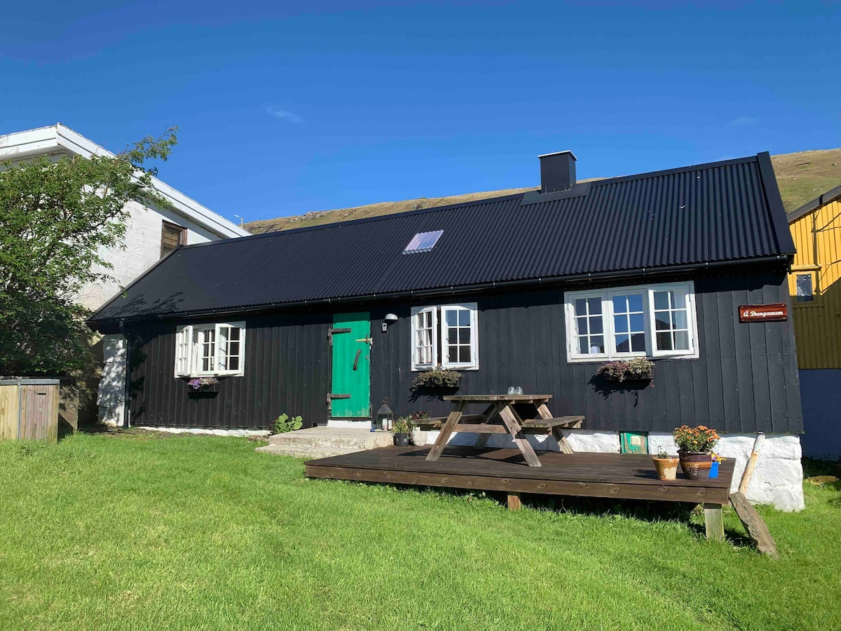 A charming, traditional house features a dark exterior complemented by a green door. Outdoor seating is provided on a wooden terrace, surrounded by a well-manicured lawn. Flower boxes adorn the windows, enhancing the inviting appearance of this historic building.