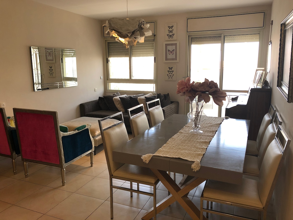 A dining area is centered around a modern table with seating for six. Plush chairs in vibrant colors contrast against neutral walls. A cozy seating arrangement with a large sofa is visible in the background, while natural light enters through expansive windows.