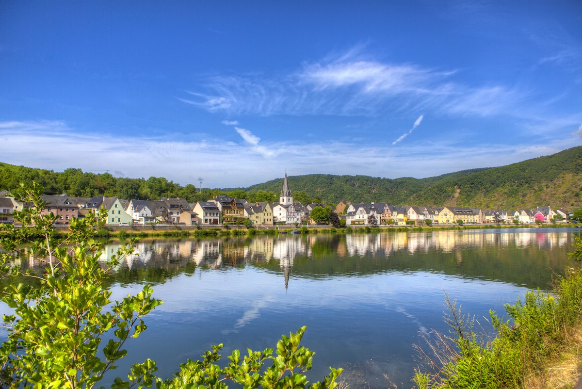 A serene riverside view showcases an array of quaint buildings along the water's edge. The reflection of the structures is visible in the calm river, framed by lush green hills and a clear blue sky, creating a peaceful natural setting.