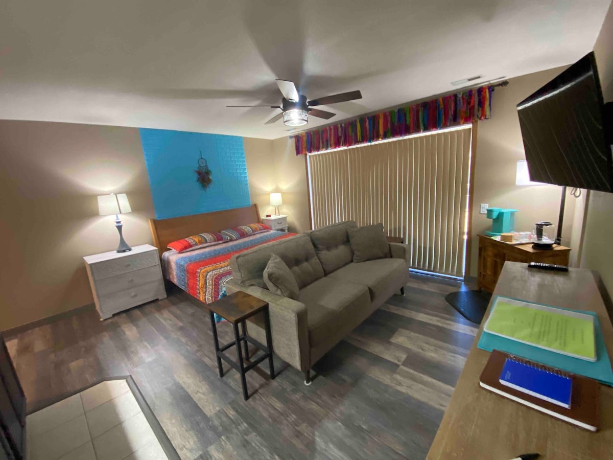 A spacious area is highlighted by a king-size bed adorned with a colorful striped blanket. A comfortable sofa faces a flat-screen TV, while a small table is nearby. Natural light enters through vertical blinds, and decorative lamps provide additional illumination.