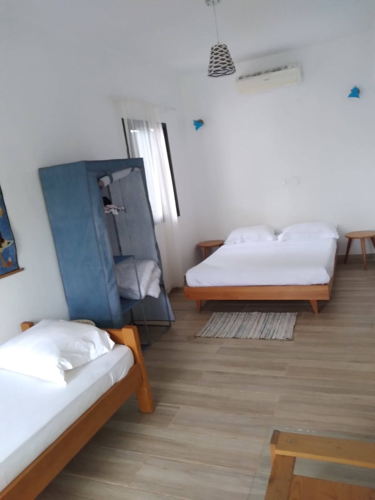 The room features three wooden beds with simple bedding, complemented by a closet with hanging space. Light-colored wooden flooring creates a spacious feel, while natural light enters through the window with a light curtain. Wall art adds subtle color and interest to the room.