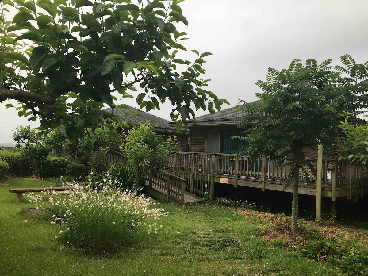 A wooden house is surrounded by greenery, featuring a deck that leads up to the entrance. Flowering plants and trees are positioned in the foreground, while a spacious lawn offers a serene outdoor space.