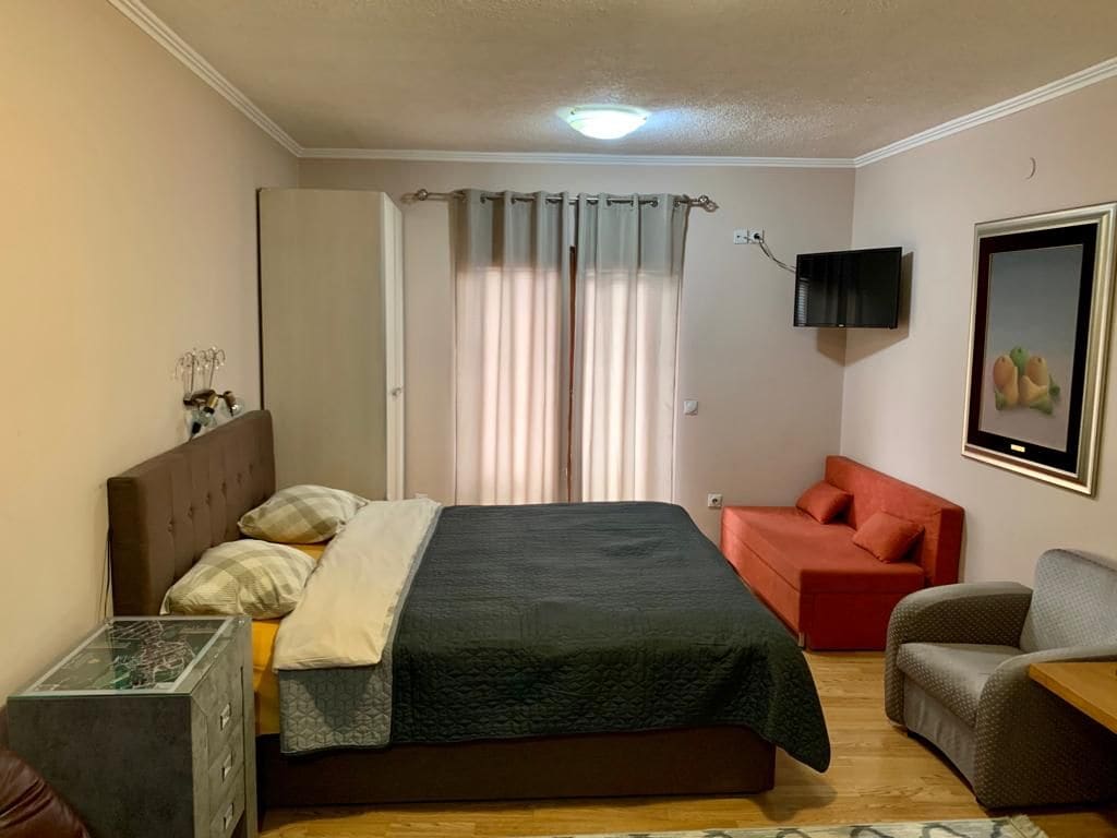 A spacious sleeping area features a large bed with neutral linens and decorative pillows. A small red sofa is positioned against the wall, while a comfortable armchair complements the space. Natural light filters through sheer curtains, highlighting the warm wood flooring.