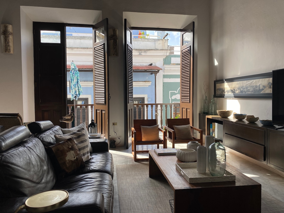 Natural light fills the living area through open shutters, illuminating a modern seating arrangement. A stylish wooden coffee table sits at the center, complemented by artistic decor. The balcony overlooks Calle Sol, enhancing the apartment's connection to the vibrant surroundings.