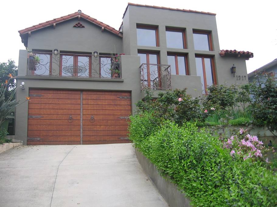 A modern two-story house features a textured exterior painted in neutral tones. Large windows, adorned with decorative ironwork, illuminate the living space. The front driveway is flanked by lush greenery and colorful flowers, leading up to a double wooden garage door.