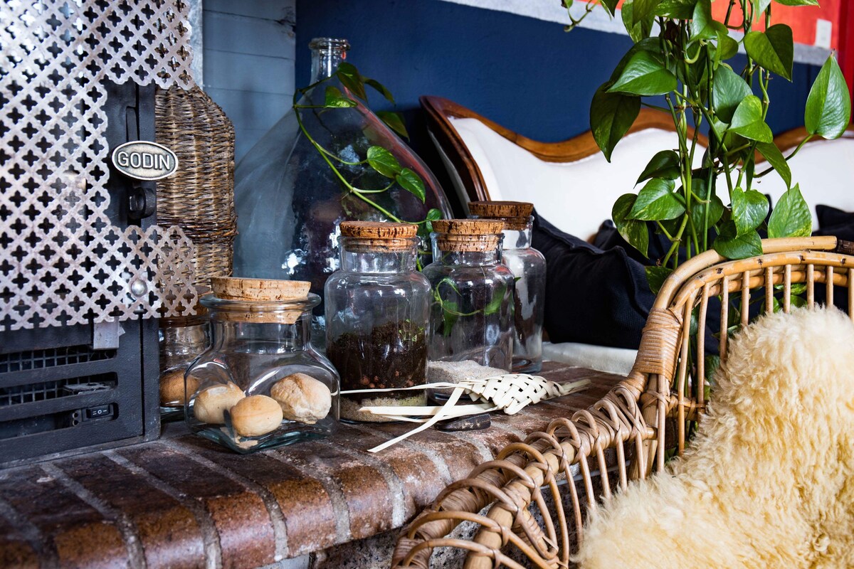 A rustic seating area features a woven chair adorned with a soft throw. A collection of jars, some filled with soil, are displayed on a dark brick ledge near a vintage-styled heater, accompanied by green plants adding a natural touch.