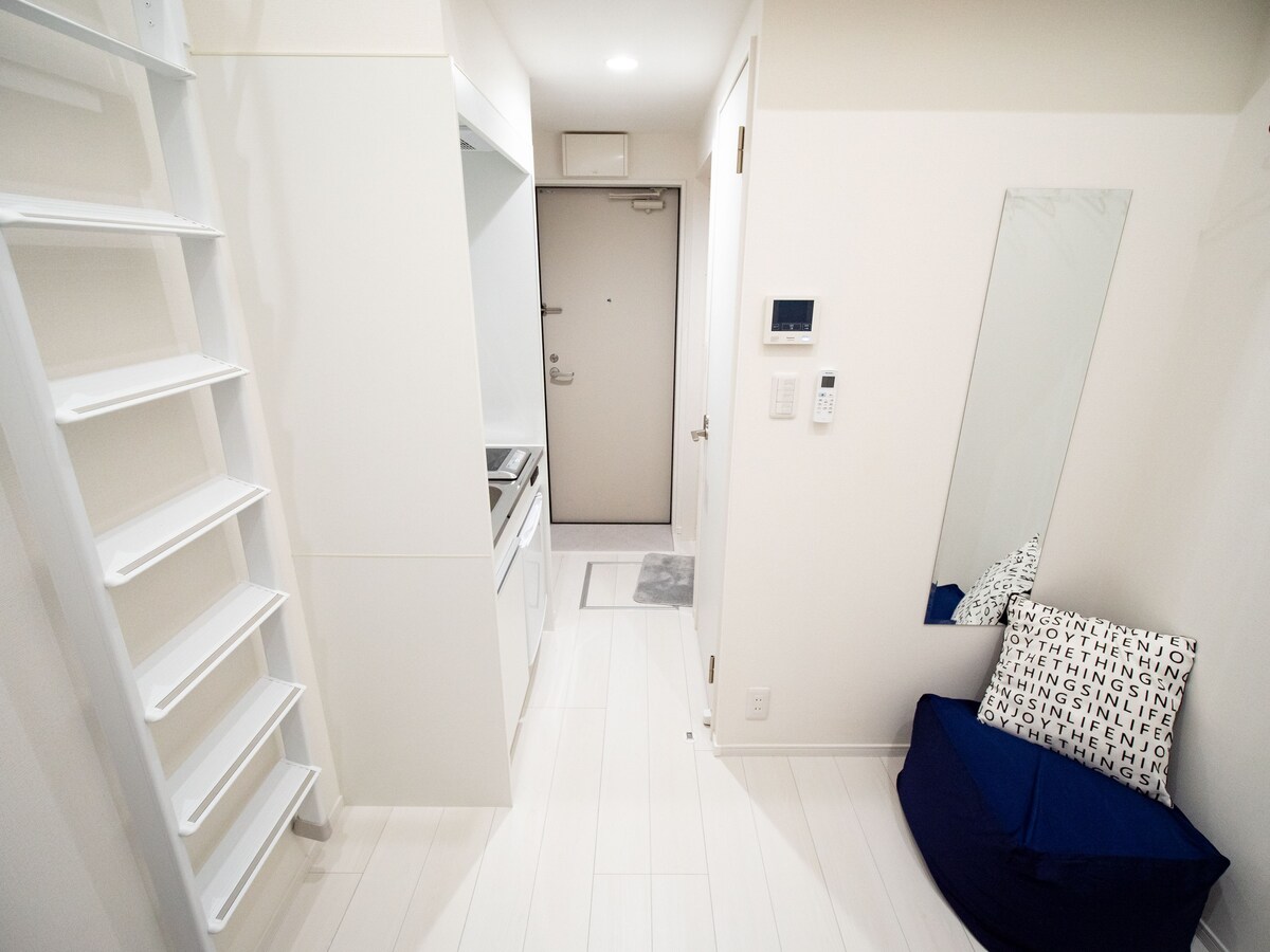 A bright hallway connects the living space to the entrance. White walls and flooring create an airy ambiance. A compact kitchen is visible on the left, with a mirror and a cozy seating area on the right, featuring a blue cushion with patterned pillows.