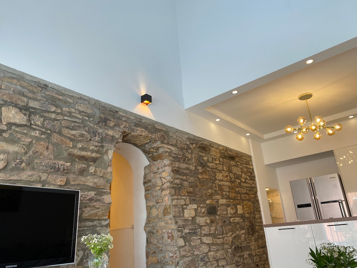 The interior features a blend of modern and historic elements, highlighted by a stone wall that adds texture. A contemporary light fixture casts warm illumination over the space. In the background, the modern kitchen is visible with stainless steel appliances, creating a sense of openness.