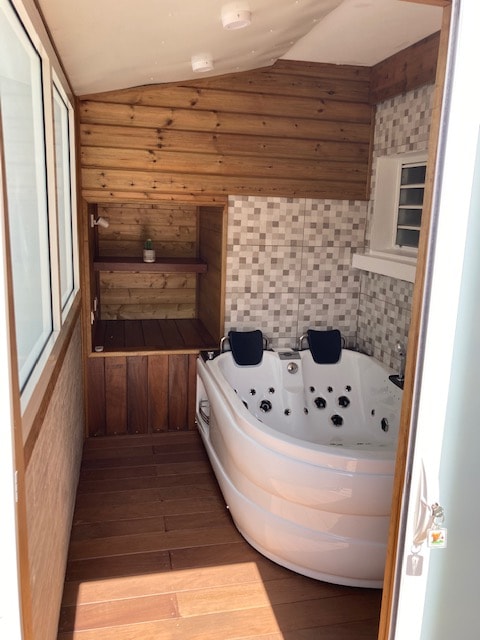 A private space is featured with a modern hydrotherapy tub, surrounded by warm wooden walls and a tiled accent wall. Natural light enters through large windows, illuminating the relaxing atmosphere. A wooden shelf is placed nearby for convenience, enhancing the overall tranquility.