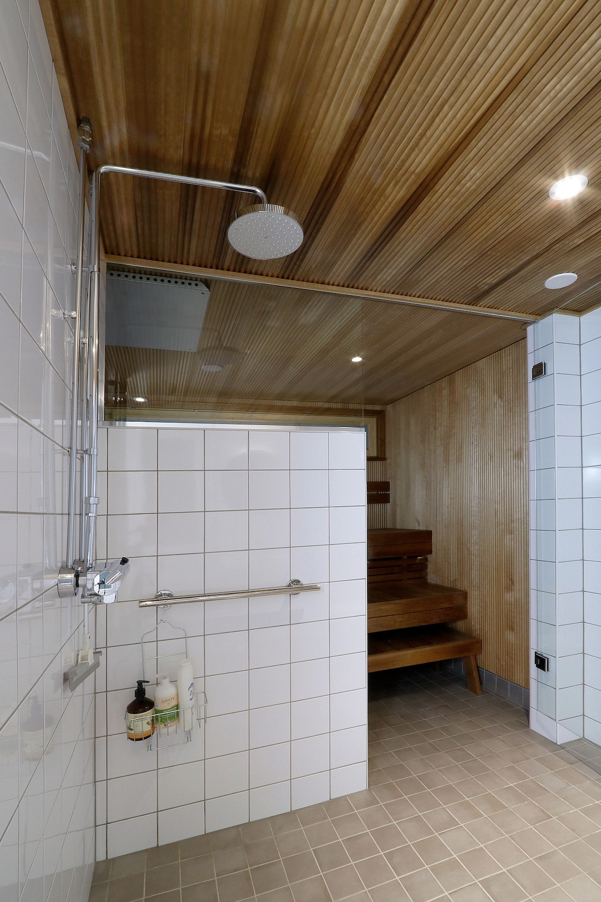 A clean and modern bathroom is presented, featuring a spacious shower area with a rain showerhead. Wooden accents are observed on the ceiling and seating. The tiled walls and floor provide a bright, simple aesthetic. Bath products are conveniently arranged on the wall-mounted shelf.