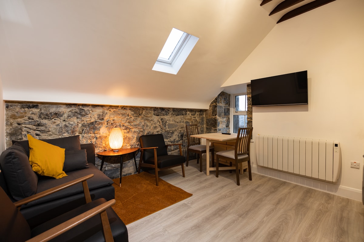 A cozy living area is presented with a comfortable sofa and chairs arranged around a small table. The space features a skylight that allows natural light to fill the room, highlighting the stone wall. A modern television is mounted on the wall.
