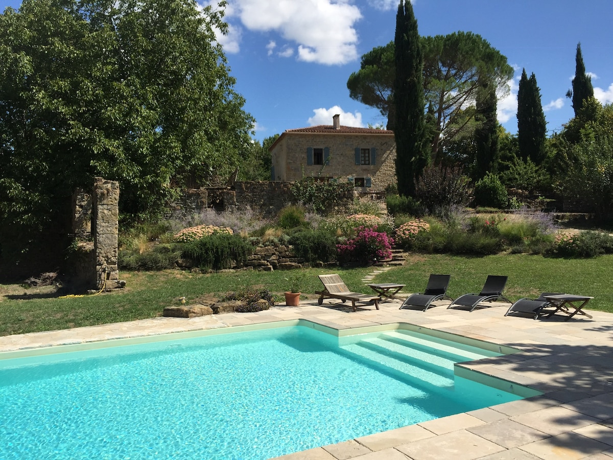 A serene pool area features clear turquoise water, surrounded by a spacious stone patio. Several lounge chairs are arranged nearby, inviting relaxation under the sun. Lush greenery, flowering plants, and tall trees enhance the tranquil landscape, with the farmhouse visible in the background.
