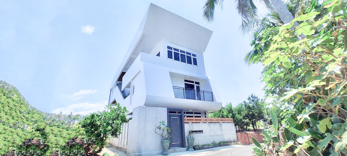 A modern three-story building features clean lines and a minimalist design. Large windows and a balcony provide natural light and outdoor access. Surrounding greenery adds a touch of nature, enhancing the serene setting, while the well-maintained exterior suggests a welcoming atmosphere.