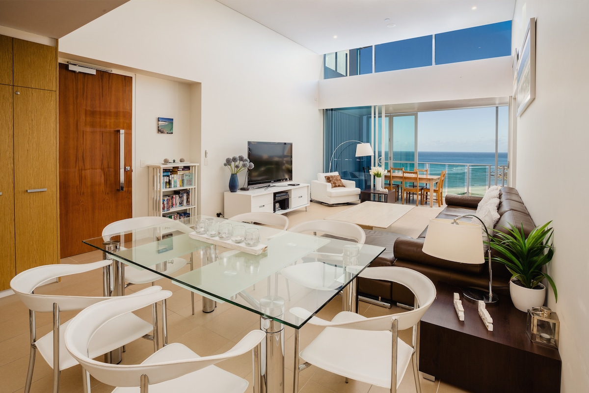 An open-plan living area is highlighted, featuring a glass dining table surrounded by white chairs. A brown leather sofa is positioned across from a television. Large windows provide a view of the ocean, with natural light illuminating the space.