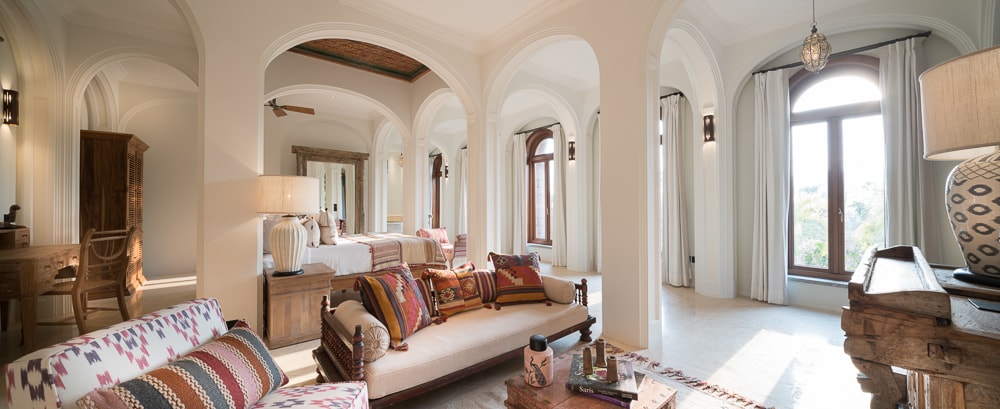 The spacious living area showcases arched windows allowing natural light to fill the room. Comfortable seating is arranged around a coffee table, with handcrafted furnishings and vibrant textiles adding character. Two elegantly designed beds are positioned in the background, creating a warm and inviting space.