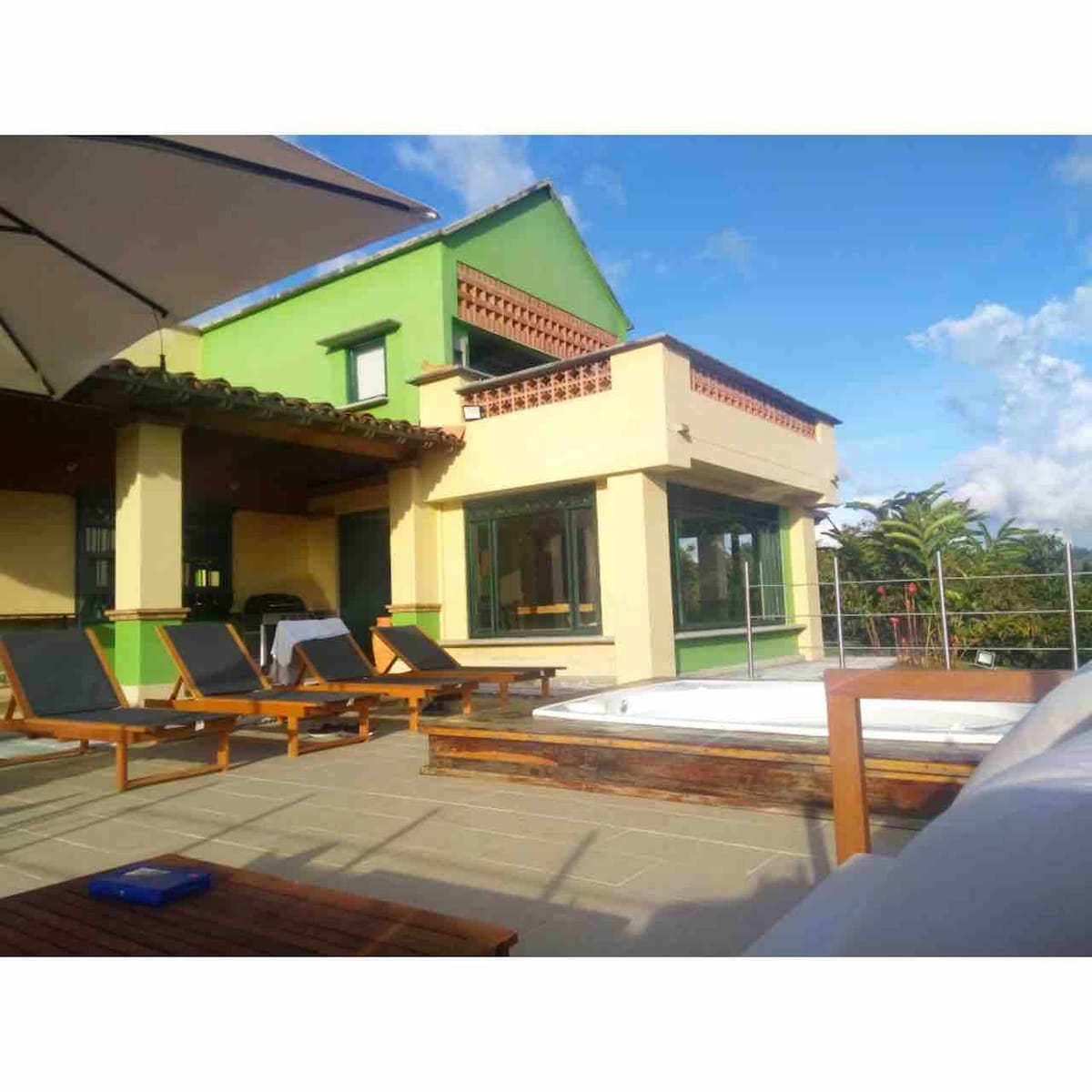 A spacious outdoor area features loungers positioned under an umbrella, along with a jacuzzi for eight people. The colorful house, showing a green facade, complements the clear blue sky. A well-lit patio area offers a relaxing space for guests.