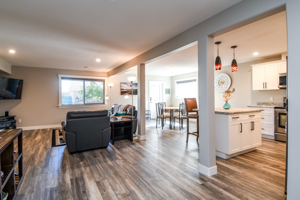 An open-concept living space is visible, featuring a comfortable seating area with a dark sofa and a television mounted on the wall. Light wood flooring extends throughout. A dining table with chairs is positioned nearby, while a modern kitchen with white cabinetry is seen in the background.
