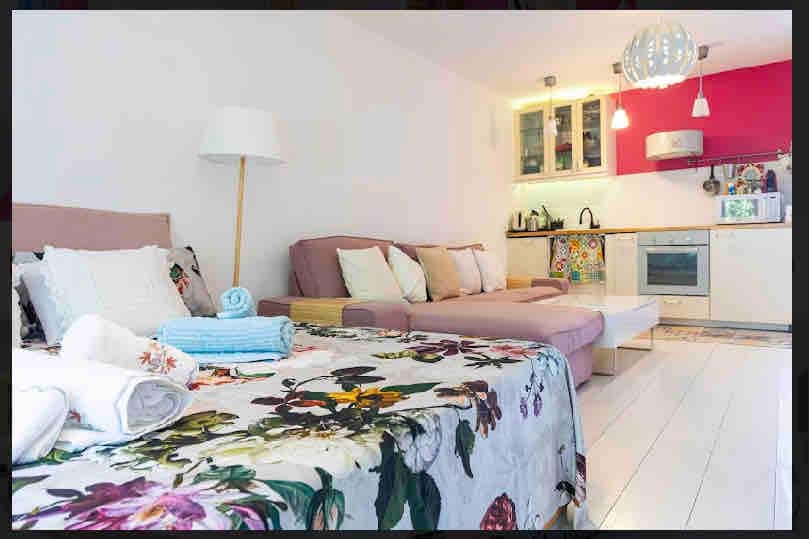 A bright interior space features a spacious open layout. A floral bedspread covers a bed, accompanied by a neatly arranged stack of towels. A modern kitchenette, equipped with appliances, stands against a vibrant red wall. Natural light illuminates the area, enhancing the warm atmosphere.
