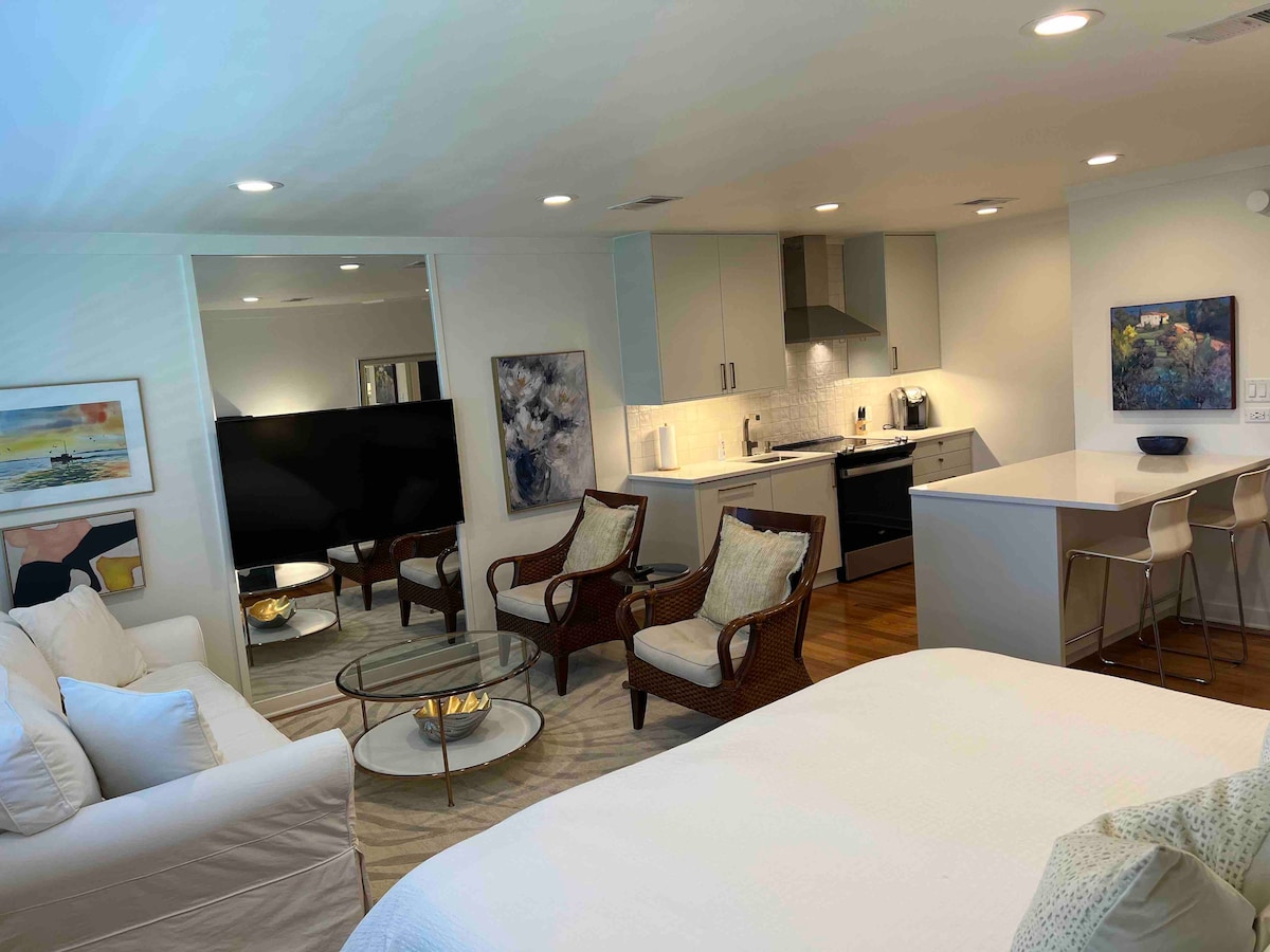 A modern studio space is presented, featuring a spacious bed and a cozy seating area with two armchairs. A flat-screen television is positioned across from the seating. The kitchen area includes sleek cabinets and bar seating at the counter, with subtle artwork adorning the walls.