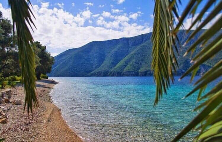 A tranquil shoreline extends along the crystal-clear waters of the Ionian Sea, framed by lush greenery and distant mountains. The sandy beach curves gently, inviting relaxation and exploration. The bright sky features soft clouds, enhancing the natural beauty of the scene.